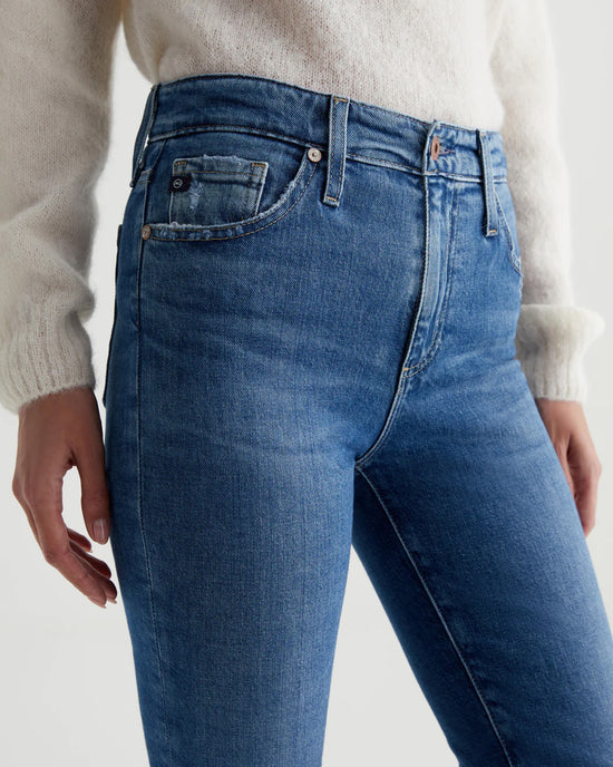 Close-up of a person wearing AG Jeans Mari Crop in Alibi Destructed with a hand partially inserted into a pocket, showing details like buttons and stitching.
