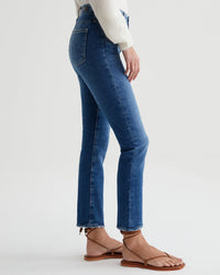 Side view of a woman wearing AG Jeans Mari Crop in Alibi Destructed high-waist straight leg jeans and brown sandals, standing against a white background.