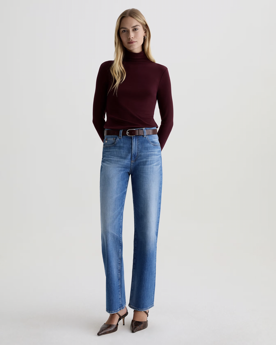 A woman stands against a plain background wearing a burgundy turtleneck, The Brinley Straight in 13Ys Allure by AG Jeans with a brown belt, and pointed brown heels. Her arms are behind her back and she has long, straight blonde hair.