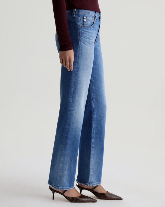 A person stands against a plain light background wearing AG Jeans' The Brinley Straight in 13Ys Allure, a long-sleeve burgundy top, and brown pointed-toe heels.