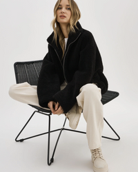 Woman in a Lamarque Kim Jacket in Black Sherpa and cream pants sitting on a black wire chair.