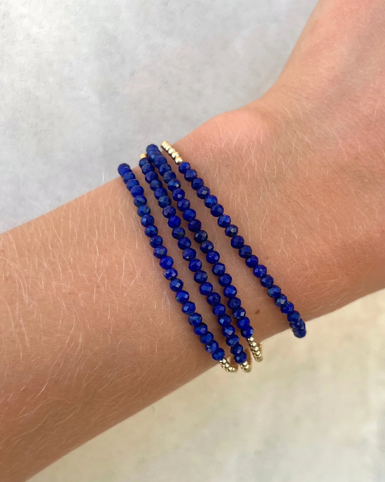 A wrist adorned with multiple Karen Lazar Design 2MM Sig Bracelets with Lapis & Yellow Gold against a neutral background, each featuring 14k Yellow Gold filled beads.