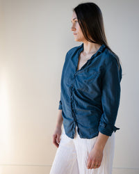 A woman with long brown hair stands sideways against a plain background, wearing the LLEA BLUE Carrie Blouse in Moonlit Ocean Cotton Silk with a deep v-neck and rolled sleeves, paired with loose white pants.