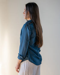 A woman with long brown hair stands sideways, facing left, wearing the LLEA BLUE Carrie Blouse in Moonlit Ocean Cotton Silk and light-colored pants against a neutral background.