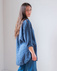 A woman with long brown hair wears the LLEA BLUE Ciel Tunic Blouse in Moonlit Ocean Linen and blue jeans, standing sideways and glancing over her shoulder in a minimalist, light-filled room with white walls.