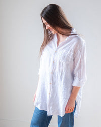 A woman with long brown hair wears the LLEA BLUE Ciel Tunic Blouse in White Linen and blue jeans, standing against a plain light background while looking down and smiling gently.