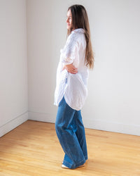 A woman with long brown hair stands sideways in a bright room, wearing the LLEA BLUE Ciel Tunic Blouse in White Linen and wide-leg blue jeans for an effortless, oversized look on a light wooden floor.