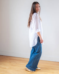 A woman with long brown hair wears the LLEA BLUE Ciel Tunic Blouse in White Linen, paired with blue wide-leg jeans and gold shoes, standing on a wooden floor against a plain white wall as she glances over her shoulder.