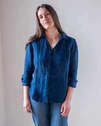 A person with long brown hair stands against a plain light wall, wearing the LLEA BLUE Carrie Fitted Blouse in Indigo and blue jeans, highlighting a slim silhouette while looking calmly at the camera with relaxed posture.