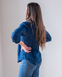 A woman with long brown hair stands sideways, hands on hips, wearing the LLEA BLUE Carrie Fitted Blouse in Indigo and blue jeans, facing a light-colored wall.