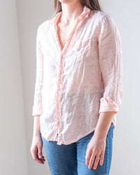 A person in the LLEA BLUE Carrie Fitted Blouse in Bermuda Sand Linen—light pink, long-sleeved with rolled-up sleeves and a deep v-neck—paired with blue jeans, stands against a light background, head partially out of frame.