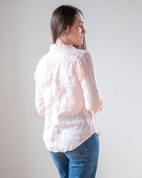 A person with long brown hair wears the LLEA BLUE Carrie Fitted Blouse in Bermuda Sand Linen and blue jeans, standing slightly turned away against a plain light background.