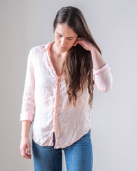 A woman with long brown hair, wearing an LLEA BLUE Carrie Fitted Blouse in Bermuda Sand Linen and blue jeans, stands against a plain background, looking down and touching her hair with one hand.