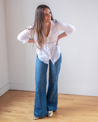A woman with long brown hair stands indoors on a wooden floor, smiling and leaning forward with her hands on her hips. She wears the LLEA BLUE Carrie Fitted Blouse in White Linen, blue wide-leg jeans, and gold shoes.