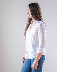A woman with long brown hair wears the LLEA BLUE Carrie Fitted Blouse in White Linen and blue jeans, standing against a plain light background, facing right in a relaxed pose.