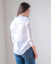 A woman with long brown hair, wearing the LLEA BLUE Carrie Fitted Blouse in White Linen and blue jeans, stands turned slightly away against a plain light background.