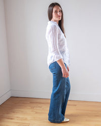 A woman with long brown hair poses sideways on a wooden floor, wearing the LLEA BLUE Carrie Fitted Blouse in White Linen and wide-leg blue jeans, looking at the camera with a neutral expression against a plain white wall.