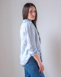 A woman with long brown hair wears the LLEA BLUE Carrie Fitted Blouse in Aqua Chambray Linen and blue jeans, standing sideways and looking over her shoulder against a plain light background.