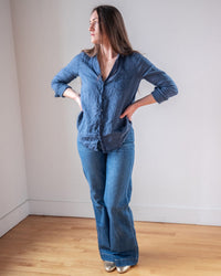 A person with long hair stands indoors on a wooden floor, wearing LLEA BLUE’s Carrie Fitted Blouse in Moonlit Ocean Linen and flared jeans. With hands on hips, they glance sideways, highlighting the blouse's relaxed elegance.