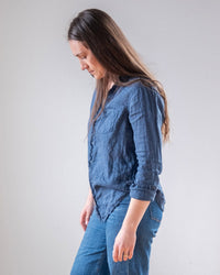 A woman with long brown hair, wearing the LLEA BLUE Carrie Fitted Blouse in Moonlit Ocean Linen and blue jeans, stands in profile looking down against a plain light background.
