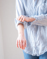 A person wears the LLEA BLUE Alix Top in Aqua Chambray Linen, rolling up their left sleeve. Only their torso and arms are shown against a plain, light background.