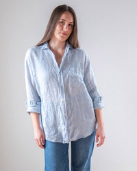 A person with long brown hair wears the LLEA BLUE Alix Top in Aqua Chambray Linen, a loose-fitting linen button-down with rolled-up sleeves, paired with blue jeans, standing against a plain light background.