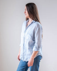 A woman with long brown hair, wearing an LLEA BLUE Alix Top in Aqua Chambray Linen and blue jeans, stands facing left against a plain light background.