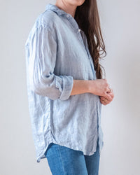 A person with long brown hair wears the LLEA BLUE Alix Top in Aqua Chambray Linen with rolled sleeves and blue jeans, standing sideways against a plain light background.