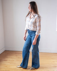 A woman with long brown hair stands indoors on a hardwood floor, wearing the LLEA BLUE Dorie Top in White, Blue & Wheat Linen Plaid, blue high-waisted wide-leg jeans, and gold shoes, looking to her right.