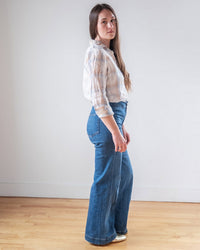 A woman with long brown hair stands sideways, looking at the camera. She wears the LLEA BLUE Dorie Top in White, Blue, & Wheat Linen Plaid with high-waisted wide-leg blue jeans, on a light wood floor against a plain white wall.