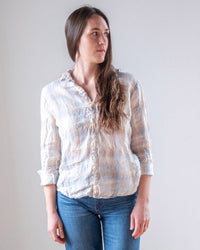 A woman with long brown hair wears the LLEA BLUE Dorie Top in White, Blue, & Wheat Linen Plaid and blue jeans, standing against a plain light background and looking to her left with a neutral expression.
