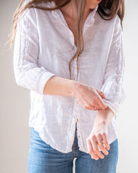 Someone in blue jeans rolls up the sleeve of their white LLEA BLUE Dorie Top in Linen. Their long brown hair partly obscures their face.