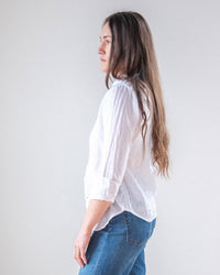 A woman with long brown hair, wearing the Dorie Top in White Linen from LLEA BLUE and blue jeans, stands in profile against a plain light gray background.