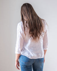 A person with long brown hair, wearing the LLEA BLUE Dorie Top in White Linen and blue jeans, stands with their back to the camera against a plain light background.
