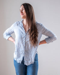 A woman with long brown hair stands with her hands on her hips, wearing the LLEA BLUE Dorie Top in Aqua Chambray Linen and jeans, looking off to the side with a slight smile against a plain background.