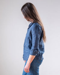 A woman with long brown hair, wearing the LLEA BLUE Dorie Top in Moonlit Ocean Linen and blue jeans, stands in profile against a plain light background.