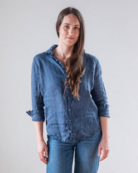 A woman with long brown hair stands against a light gray background, wearing the LLEA BLUE Dorie Top in Moonlit Ocean Linen with rolled-up sleeves and blue jeans, gazing at the camera with a neutral expression.