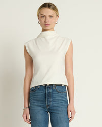 A person with blonde hair in a low bun wears the Lulu Mock Neck Top in Porcelain by Nation LTD and high-waisted blue jeans, standing against a plain light background.