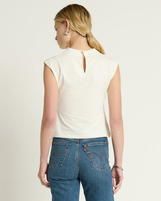 A person with long blonde hair in a low ponytail is standing and facing away, wearing the Nation LTD Lulu Mock Neck Top in Porcelain paired with high-waisted blue jeans.