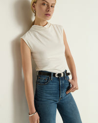 Wearing the Nation LTD Lulu Mock Neck Top in Porcelain, tucked into high-waisted blue jeans with a black belt, this person stands against a plain white wall.