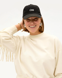 Woman smiling while wearing an adjustable Clare V. Emb Ciao Baseball Hat in Black w/ Cream and a fringed sweatshirt.