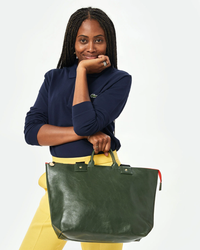 Woman holding a Clare V. Le Zip Sac in Loden Rustic tote bag, wearing a navy polo shirt and yellow trousers.