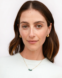A woman with a subtle smile wearing gold hoop earrings and a Clare V. Jewelry Letter Bead Necklace in Evergreen/Gold, dressed in a white sweater against a neutral background.
