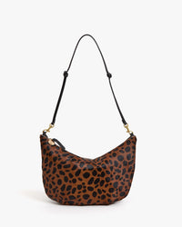 The Clare V. Lune - Tan Leopard bag has a crescent shape, tan and black leopard print hair-on leather, a black adjustable strap with gold hardware, and is shown against a plain white background.
