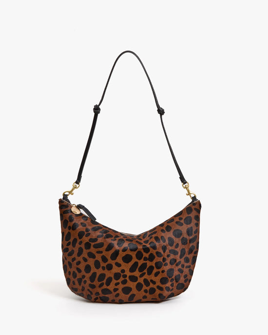 The Clare V. Lune - Tan Leopard bag has a crescent shape, tan and black leopard print hair-on leather, a black adjustable strap with gold hardware, and is shown against a plain white background.