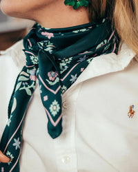 A person wears a white collared shirt, a green flower earring, and the Maison Lecomte Flament Secret Garden 100% Silk in Vert scarf styled as a square bandana around their neck.