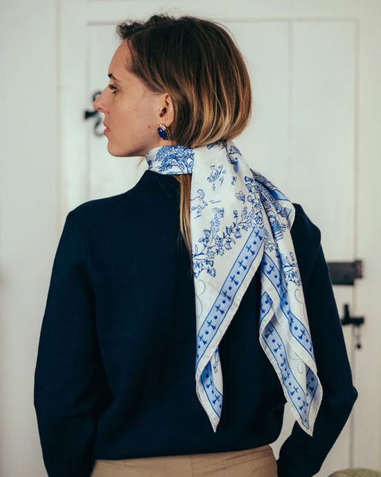A woman with light brown hair, in a navy sweater and beige pants, stands with her back to the camera. She wears Maison Lecomte Flament’s Toile de Jouy 100% Silk in Navy scarf around her neck and faces a white door.