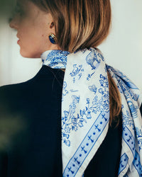A woman with blonde hair tied back wears the Maison Lecomte Flament Toile de Jouy 100% Silk in Navy scarf around her neck, paired with a navy top and blue and gold earrings. She is photographed from behind and slightly to the side.