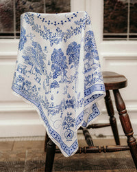 The Toile de Jouy 100% Silk in Navy scarf by Maison Lecomte Flament, featuring detailed tree, animal, and people motifs, drapes over a wooden chair in front of a glass-paneled white door, highlighting fine Italian craftsmanship.