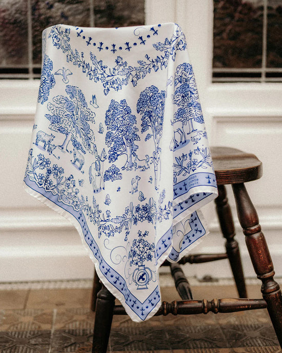 The Toile de Jouy 100% Silk in Navy scarf by Maison Lecomte Flament, featuring detailed tree, animal, and people motifs, drapes over a wooden chair in front of a glass-paneled white door, highlighting fine Italian craftsmanship.